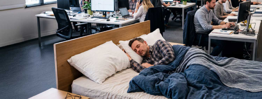 Dormire al lavoro: uomo addormentato in un letto in un open space, campagna IASO Farmacie